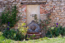 View Of A Flower Garden With A Earthenware Fountain Representing A Lion Head On An Old Brick Wall In Springtime, Piedmont, Italy 