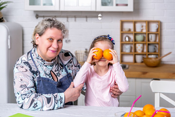 Happy little girl and her grandmother have breakfast together in a white kitchen. They are having fun and playing with fruits. Maternal care and love. Healthy eating