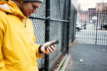 Wall Mural - Smiling man in yellow raincoat using mobile phone, standing near fence on the city street