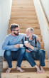 An adult hipster son and senior father sitting on stairs indoors at home, talking.