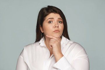 Wall Mural - Human facial expressions, feelings and emotions. Studio image of pensive thoughtful young European businesswoman with large curvy body touching her chin, thinking over creative business ideas