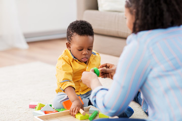 Wall Mural - childhood, kids and people concept - african american mother and her baby son playing together with wooden toy blocks kit on floor at home