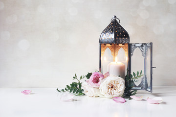 Festive greeting card, invitation for Muslim holiday Ramadan Kareem.Vintage Moroccan lantern with glowing candle, green branches, pink roses flowers and floral petals on table. Glittering bokeh lights