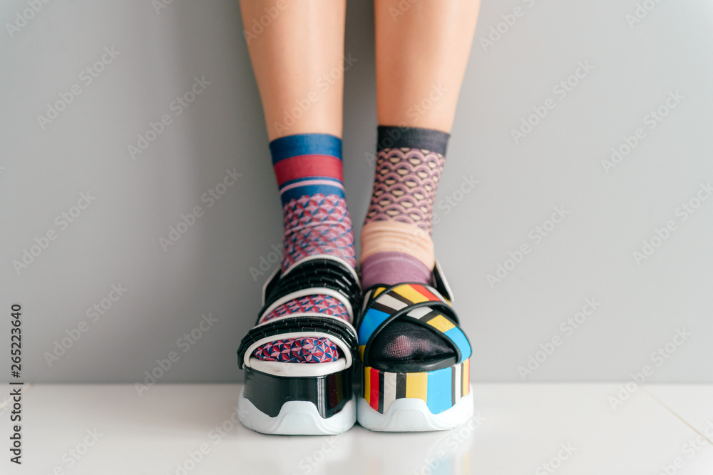 Beautiful female legs in mismatched trendy socks standing in two different fashionable high wedge leather sandals on white surface. Odd disargonized  young girl wearing high sole summer stylish shoes. - obrazy, fototapety, plakaty 