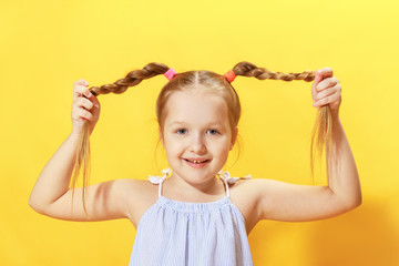 Closeup portrait of a cheerful little girl on a yellow background. The child looks into the camera and holds his hands pigtails out of hair.