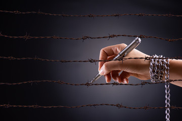 Woman hand with pen tied with chain and rusty sharp bare wire, depicting the idea of freedom of the press or expression. World press freedom and international human rights day concept.