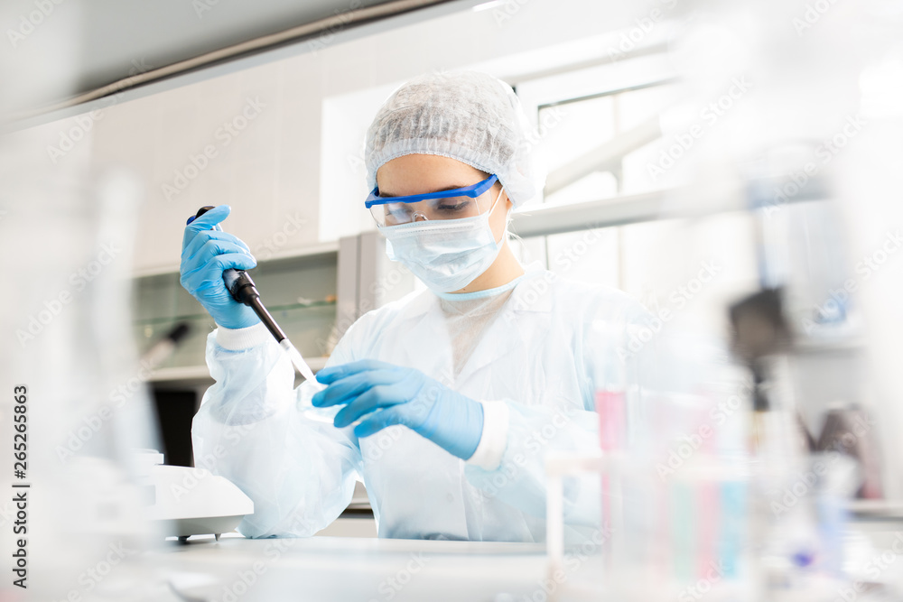 Serious concentrated female microbiologist in sterile clothing and safety goggles sitting at table and dropping reagent in petri dish while doing research in laboratory - obrazy, fototapety, plakaty 