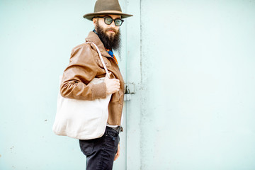 Wall Mural - Portrait of a stylish man dressed in jacket, hat and scarf standing with bag on the turquoise background outdoors