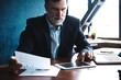 © opolja - Serious mature financial advisor sitting at the table and checking financial report using digital tablet at office.