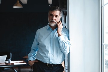 Mature businessman talking on a mobile phone standing by the window with view on city.