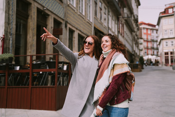 Two traveler women are walking down a street in the city. They are enjoying their company getting to know the place. One of them is pointing at a place while they are smiling. Travel concept