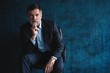 © opolja - Horizontal portrait of attractive serious mature man wearing stylish formal clothing sitting in armchair and looking at camera isolated over dark blue background.