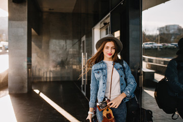 Cute pretty girl in hat with lovely face expression posing outside while wind playing with her hair before shopping. Charming female photographer in denim outfit standing on the street holding camera