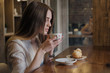© spaskov - Young beautiful brunette girl in a loft style cafe sits at a table by the window, drinks coffee and eats a dessert cake.