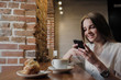 © spaskov - A young beautiful brunette girl in a loft-style cafe sits at a table by the window, drinks coffee and uses a smartphone, calls, takes pictures.