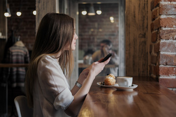 A young beautiful brunette girl in a loft-style cafe sits at a table by the window, drinks coffee and uses a smartphone, calls, takes pictures.