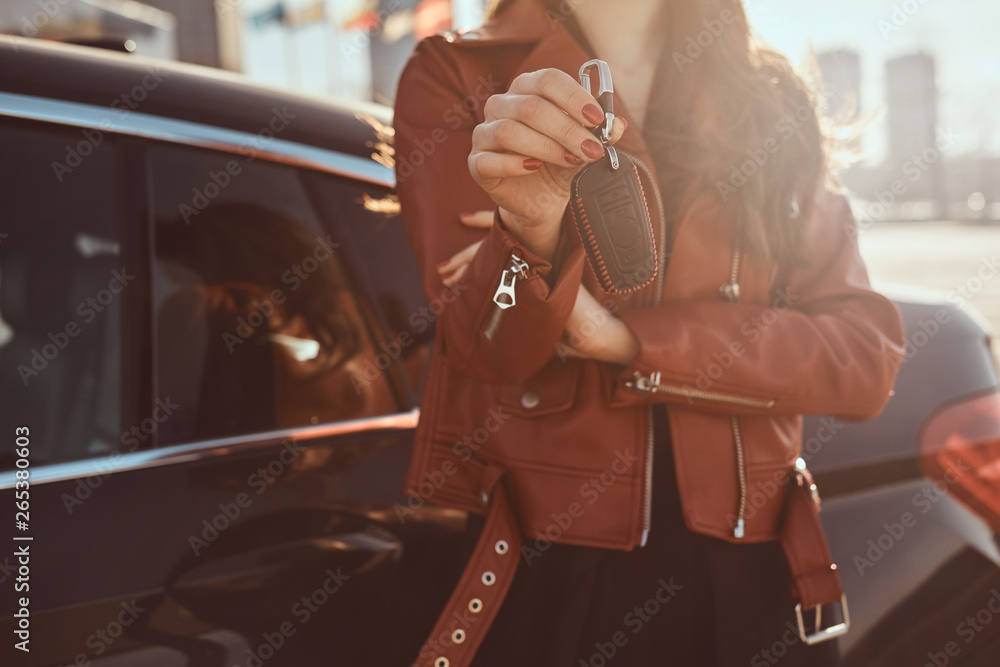 Women in red leather jacket just sold her car, which is behide her. She holds key. There are sunny day. - obrazy, fototapety, plakaty 