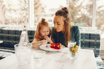 Wall Mural - young and stylish mother with long hair and a green dress sitting with her little cute daughter in the summer cafe and she feeds her daughter with a dessert