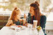 © hetmanstock2 - young and stylish mother with long hair and a green dress sitting with her little cute daughter in the summer cafe and she feeds her daughter with a dessert