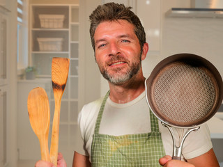 Wall Mural - young happy and attractive home cook man in apron smiling cheerful and charming holding pan and spoon enjoying domestic work and cooking hobby on modern kitchen