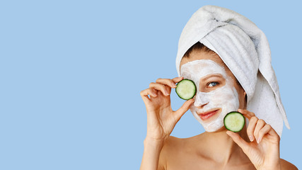 Beautiful young woman with facial mask on her face holding slices of cucumber. Skin care and treatment, spa, natural beauty and cosmetology concept.