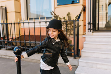 Cheerful long-haired kid holding iron tube on the street while playing after lessons under open sky. Portrait of amazing brunette little girl wearing rock style jacket and white shirt