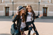 © Look! - Graceful woman with camera and leather bag looking at daughter, posing on the street in sunny day. Cheerful long-haired girl sitting on mother's knee and playfully smiling.