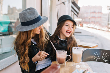 Stylish mom and pretty smiling daughter enjoying weekend together in outdoor restaurant drinking coffee and milk shake. Portrait of laughing sisters in trendy hats resting at the street cafe