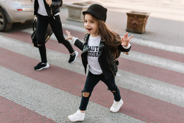 Trendy little schoolgirl walks along the pedestrian crossing after school in good mood. Portrait of joyful kid with long dark hair in cool white sneakers running on crosswalk.