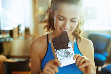 Wall Mural - smiling young sports woman in modern house biting chocolate bar