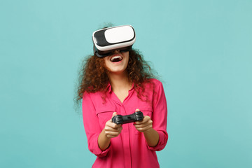 Cheerful african girl in casual clothes looking in headset, playing video game with joystick isolated on blue turquoise wall background. People sincere emotions, lifestyle concept. Mock up copy space.