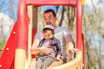 Father and toddler son sliding from children slide in the park. Child is sitting on knees of dad, father is funny and scared, kid is smiling. Dad and son spend good time outdoor