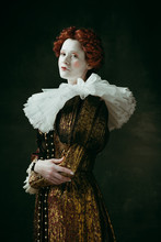 Lovely Portrait. Medieval Redhead Young Woman In Golden Vintage Clothing As A Duchess Standing Crossing Hands On Dark Green Background. Concept Of Comparison Of Eras, Modernity And Renaissance.