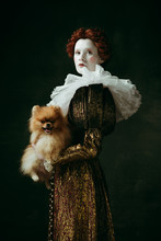 Brilliance View. Medieval Redhead Young Woman In Golden Vintage Clothing As A Duchess Holding Puppy And Standing On Dark Green Background. Concept Of Comparison Of Eras, Modernity And Renaissance.