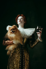 Wall Mural - Delicious. Medieval redhead young woman in golden vintage clothing as a duchess holding puppy and glass with red wine on dark green background. Concept of comparison of eras, modernity and renaissance