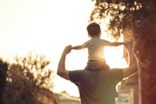 Happy Family: Young Father With His Little Child Sitting On Father's Shoulders In Summer In City At Beautiful Sunset  