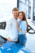 © mad_production - They dreamed of this day. Vertical shot of a handsome man hugging his happy girlfriend smiling cheerfully while touching a new car at the dealership