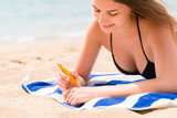 Fototapeta  - Young woman is lying on the towel at the beach and applying sunbclock from the tube on her hand