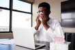 © Monkey Business - Young black businessman sitting at an office desk blowing his nose into a tissue