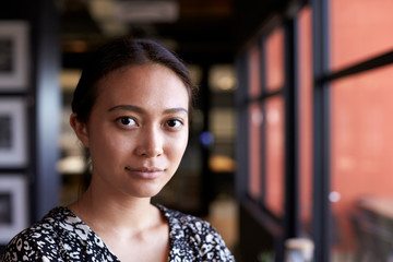 Millennial Asian businesswoman by window in office looking to camera, head and shoulders, close up