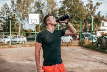 Wall Mural - Male athlete, in the summer in the city on the sports ground. In hand a shaker with water and protein. Drinks, resting after a workout fitness in nature. The concept of quenching thirst. Casual wear.