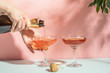 Women's hands pour champagne or wine into transparent elegant glasses. Pink pastel background bright light. Copy space.