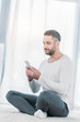 © LIGHTFIELD STUDIOS - handsome man in casual clothes sitting on carpet and using smartphone at home with sunlight