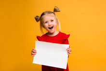 Baby Girl Holding White Sheet.Cute Little Girl With White Sheet Of Paper.yellow Background.copy Spase.Little Girl Holding Empty Sheet Of A Paper