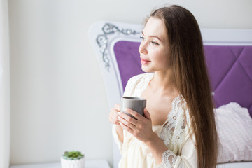 Sticker - Attractive young girl in nightwear sits on the edge of the bed by the window with a cup of coffee