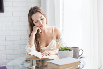 Poster - Beautiful young girl in a nightgown sits in the morning at the glass table, looks out the window and reads a book