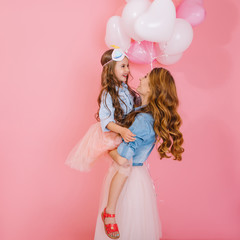 Wall Mural - Portrait of cute long-haired birthday girl with white helium balloons embracing her young curly mom after event. Mother in denim attire posing with pretty daughter at party isolated on pink background