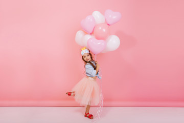 Wall Mural - Excited joyful amazing girl with long brunette hair, in princess mask on head jumping in tulle skirt with flying balloons isolated on pink background. Having fun of happy kid, expressing positivity