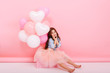 © Look! - Happy celebration of birthday party with flying balloons of charming cute little girl in tulle skirt sitting on box isolated on pink background. Expressing true positive emotions of amazing kid
