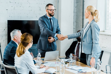 Wall Mural - handsome businessman and  blonde businesswoman shaking hands near multicultural coworkers